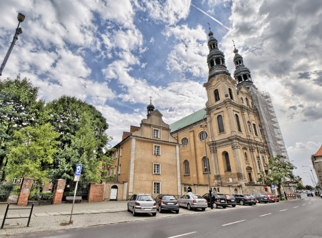 wysoki budynek z dwiema wieżami przy drodze, obok zaparkowane auta oraz ceglany mur sąsiedniego budynku