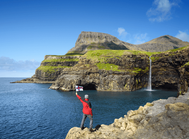 man with a Faroe Islands flag on a rocky trail, with uniquely shaped mountains and a waterfall in the background